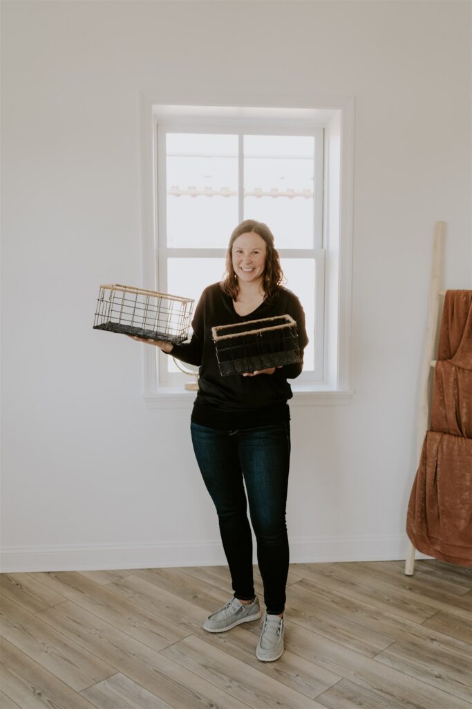 Professional organizer Bonnie standing in a bright, minimalist room with light wood floors. She is smiling broadly and holding up two black wire organization baskets with natural woven rims. In the background, a bright window sheds light on the space, and a wooden blanket ladder rests against the white wall.